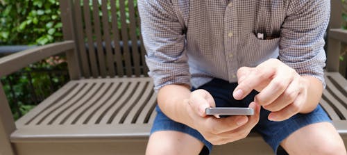 Man Wearing Brown and White Plaid Sport Shirt Sittings on Brown Bench and Using Smartphone during Day