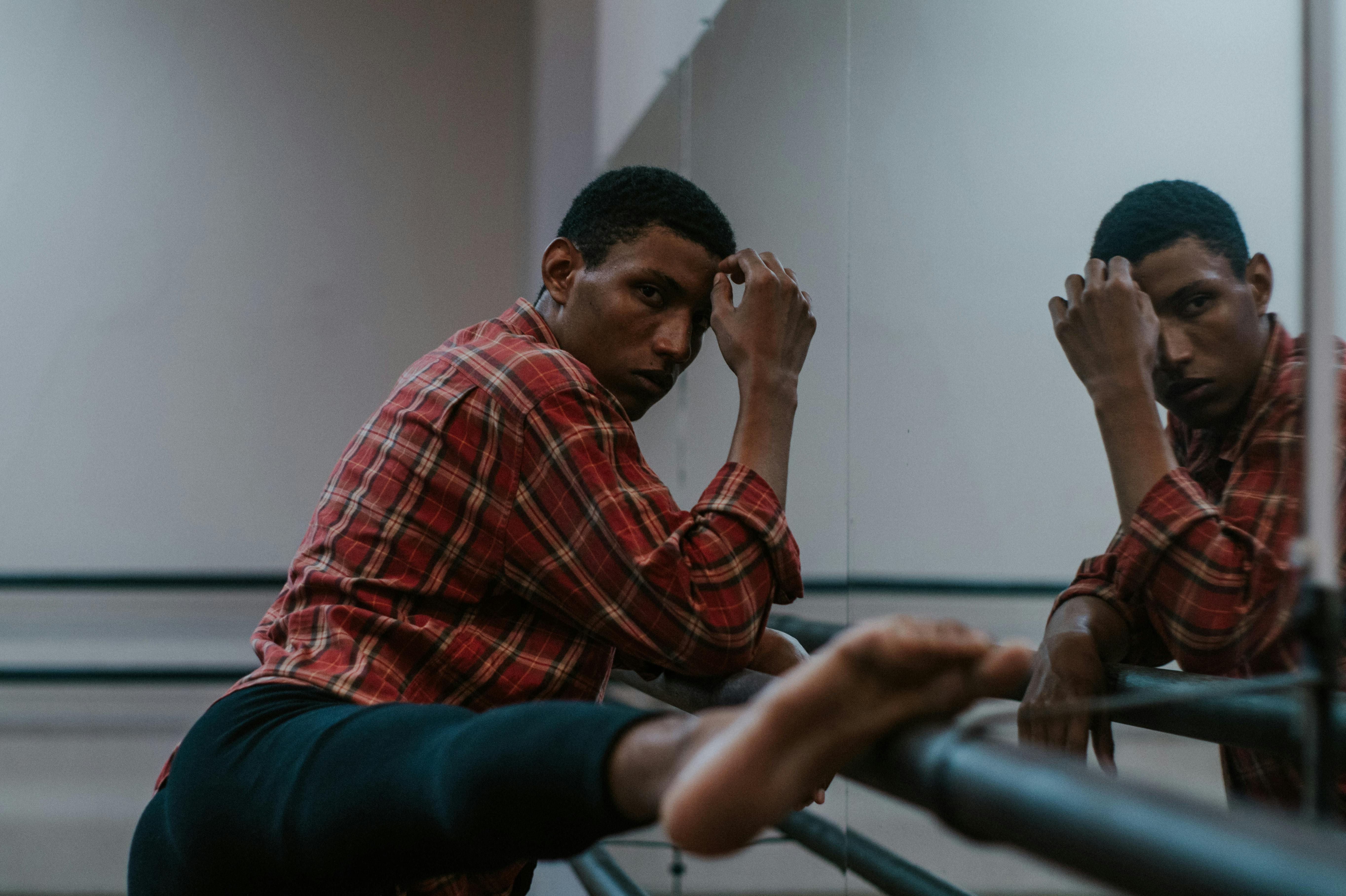 一名男性芭蕾舞演员在工作室里扶着栏杆伸展身体，镜中映出他的身影