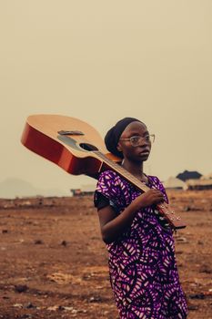Portrait of a young woman with glasses holding a guitar in an expansive outdoor setting.