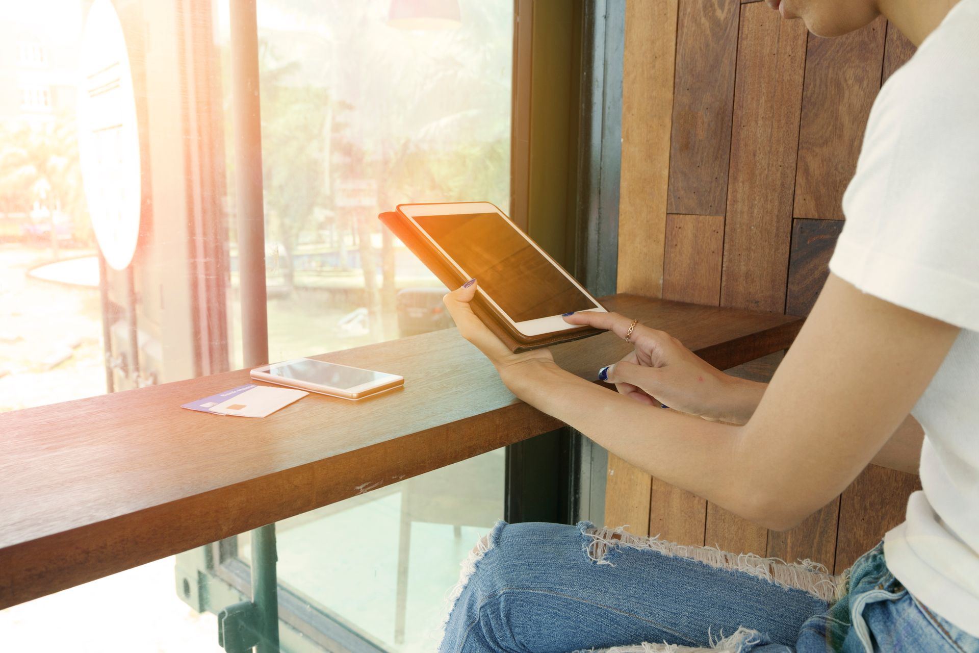 A woman uses a tablet by a sunlit cafe window, with a smartphone on the table.