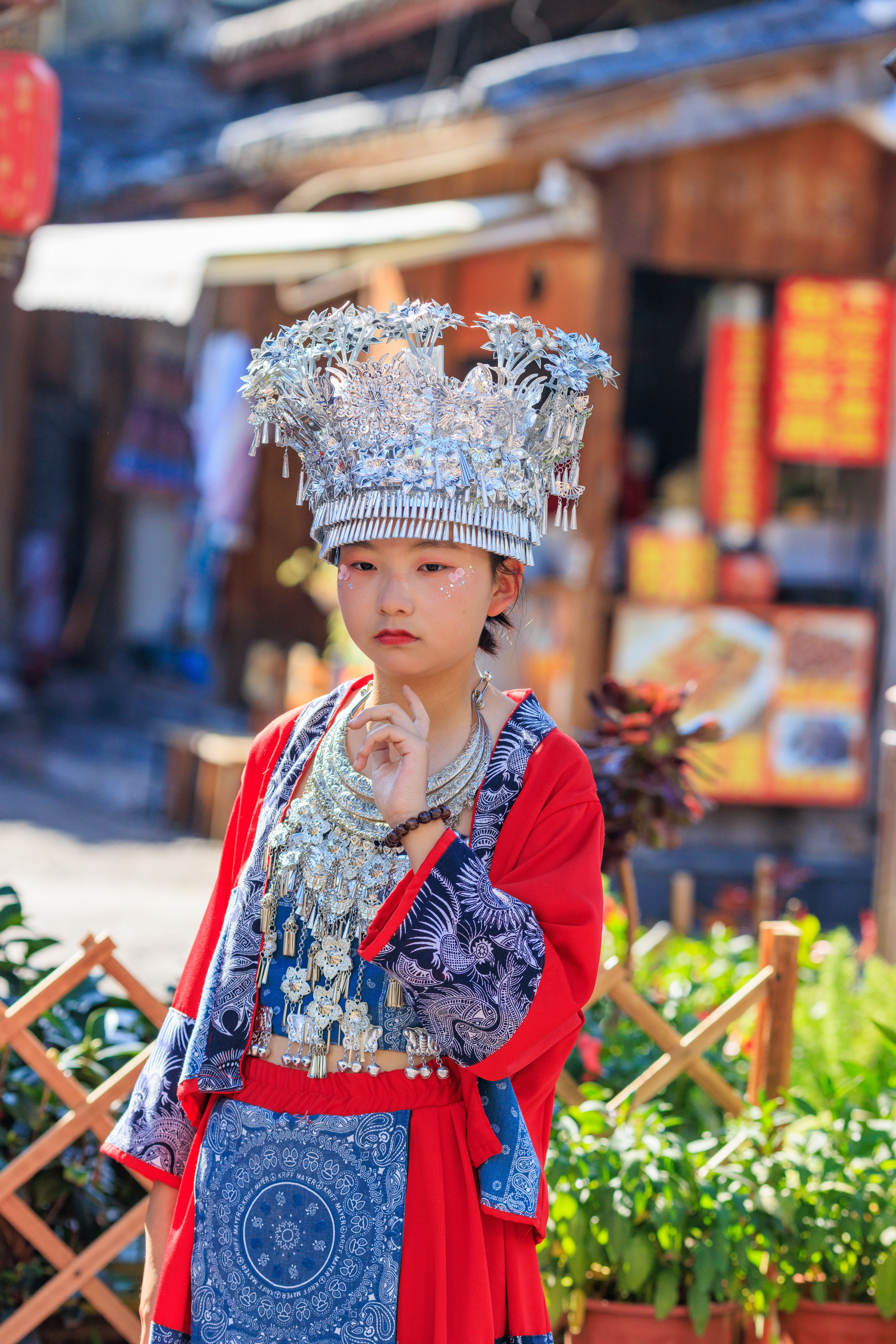 Child in traditional Miao attire with intricate silver headdress in a lively outdoor market.