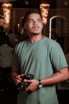 Confident young man holding camera, posing at an indoor event in warm lighting.