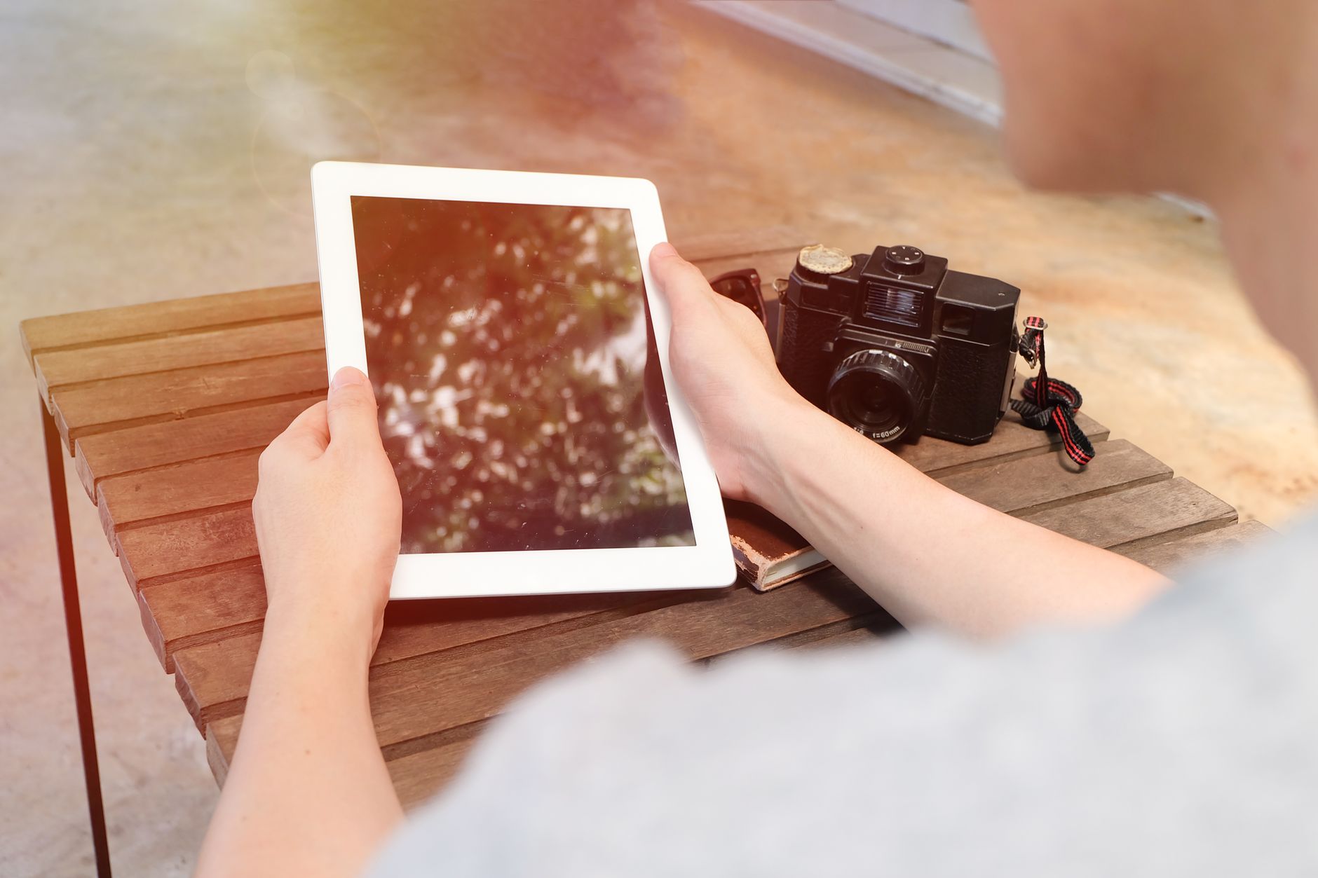 An adult holds a tablet beside a vintage camera on a wooden table indoors.