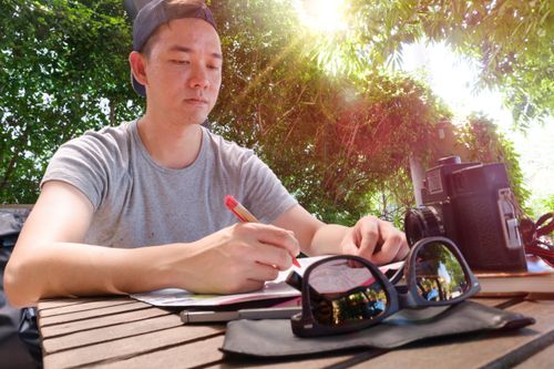 Free A man writing outdoors at a wooden table, with a vintage camera and sunglasses nearby. Stock Photo