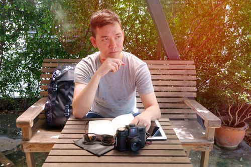 Free A man sits outdoors on a wooden bench with a vintage camera, notebook, and tablet, enjoying a sunny day. Stock Photo