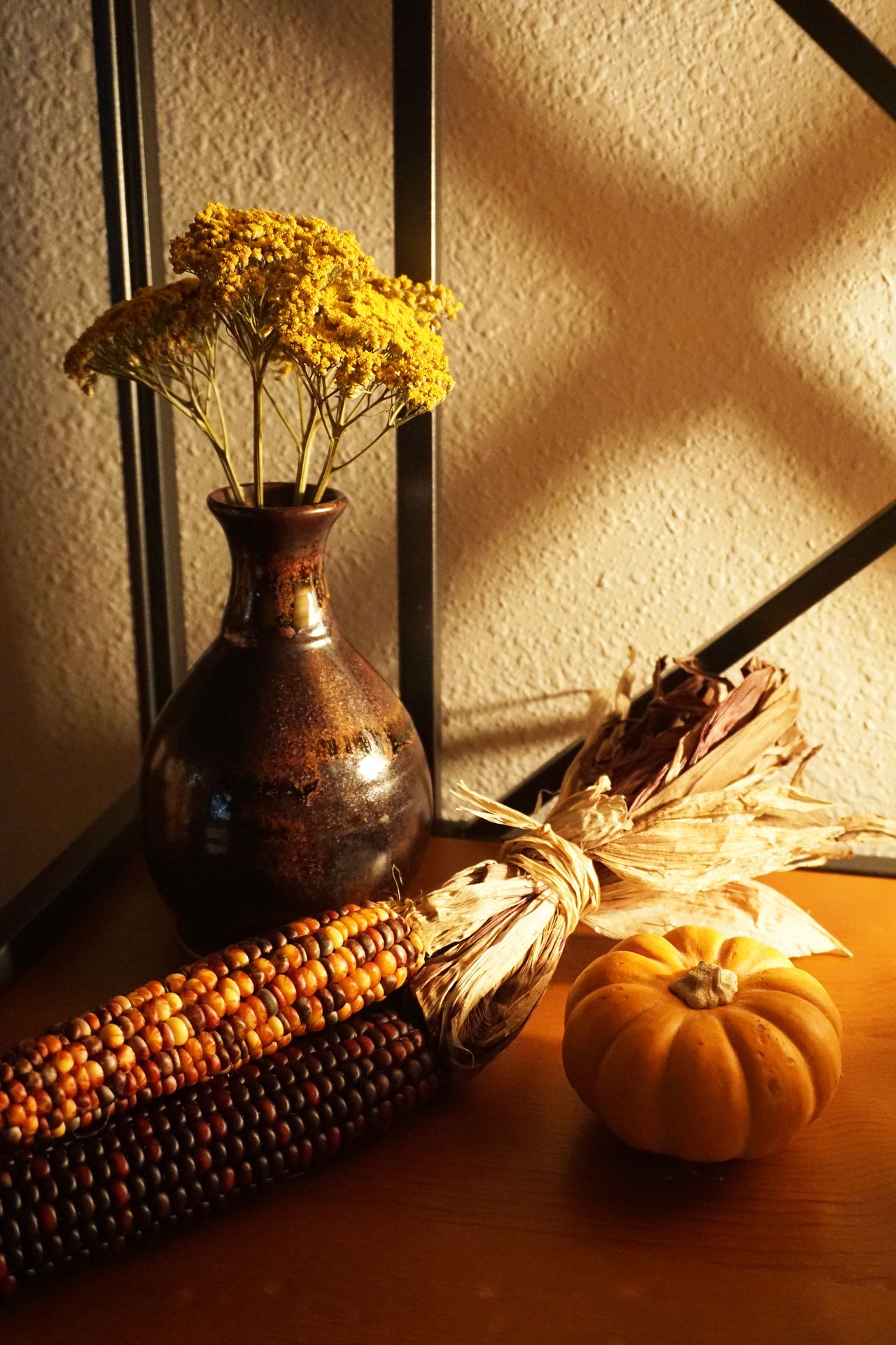 Indian corn, a mini orange pumpkin, and a vase of dried yellow flowers in warm light