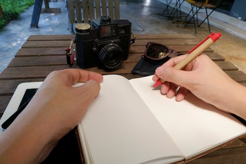 Person Writing Beside Dslr Camera on Brown Wooden Table