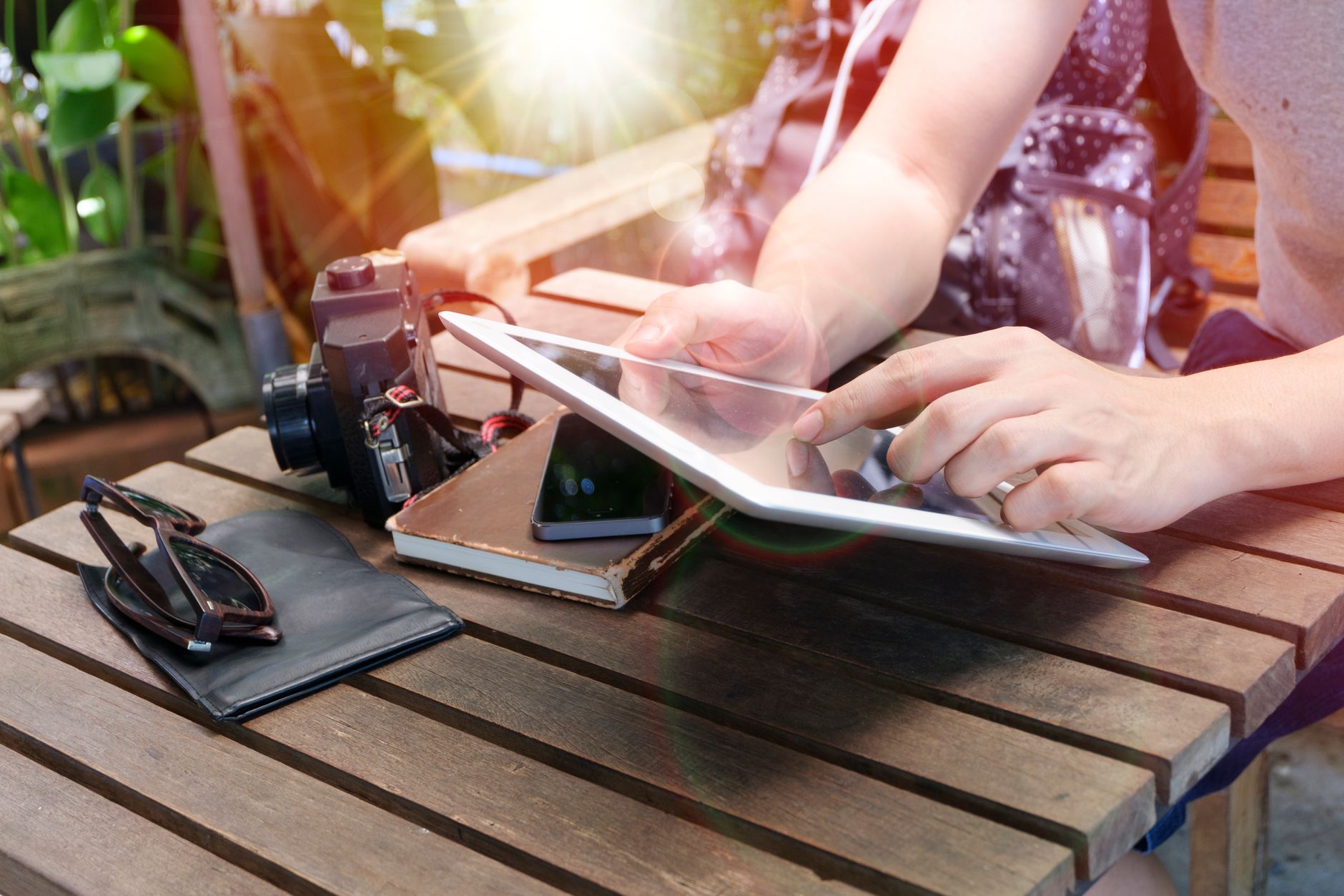Person using a tablet outdoors alongside a camera, phone, sunglasses, and notebook on a wooden table.