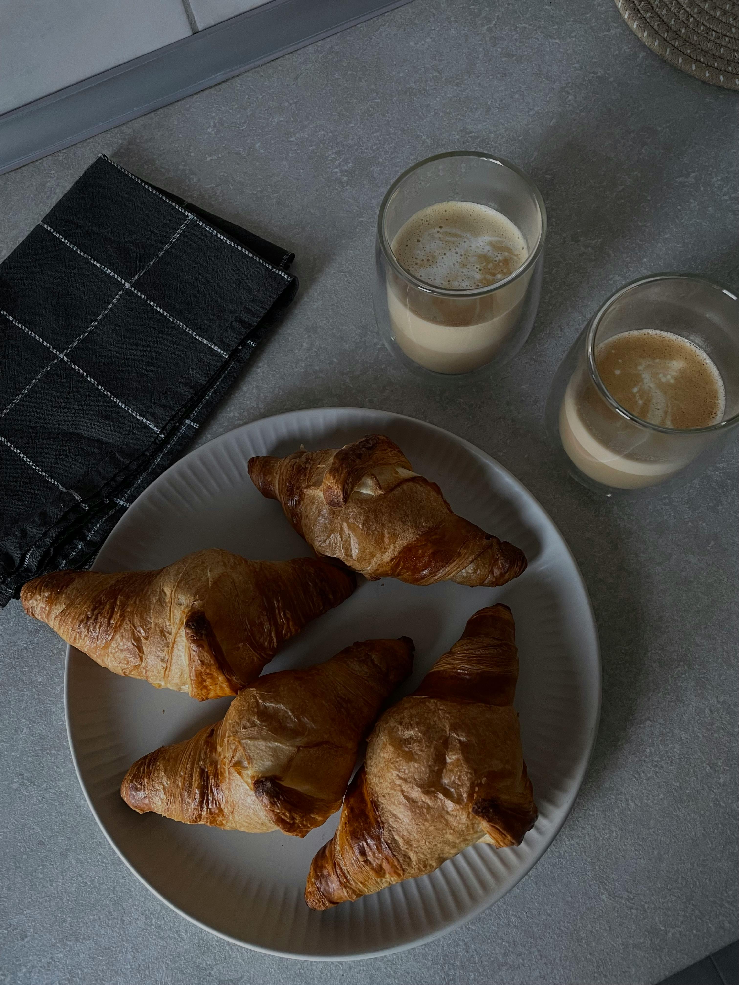 Plate of golden croissants with two glasses of coffee on a gray table setting.