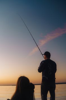 Silhouette of a man fishing at sunset by a tranquil lake with soft pastel skies.
