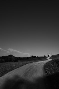 Black and white image of a winding rural road leading to a distant church, conveying solitude and tranquility.