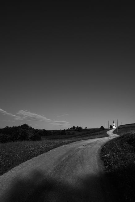 Black and white image of a winding rural road leading to a distant church, conveying solitude and tranquility.