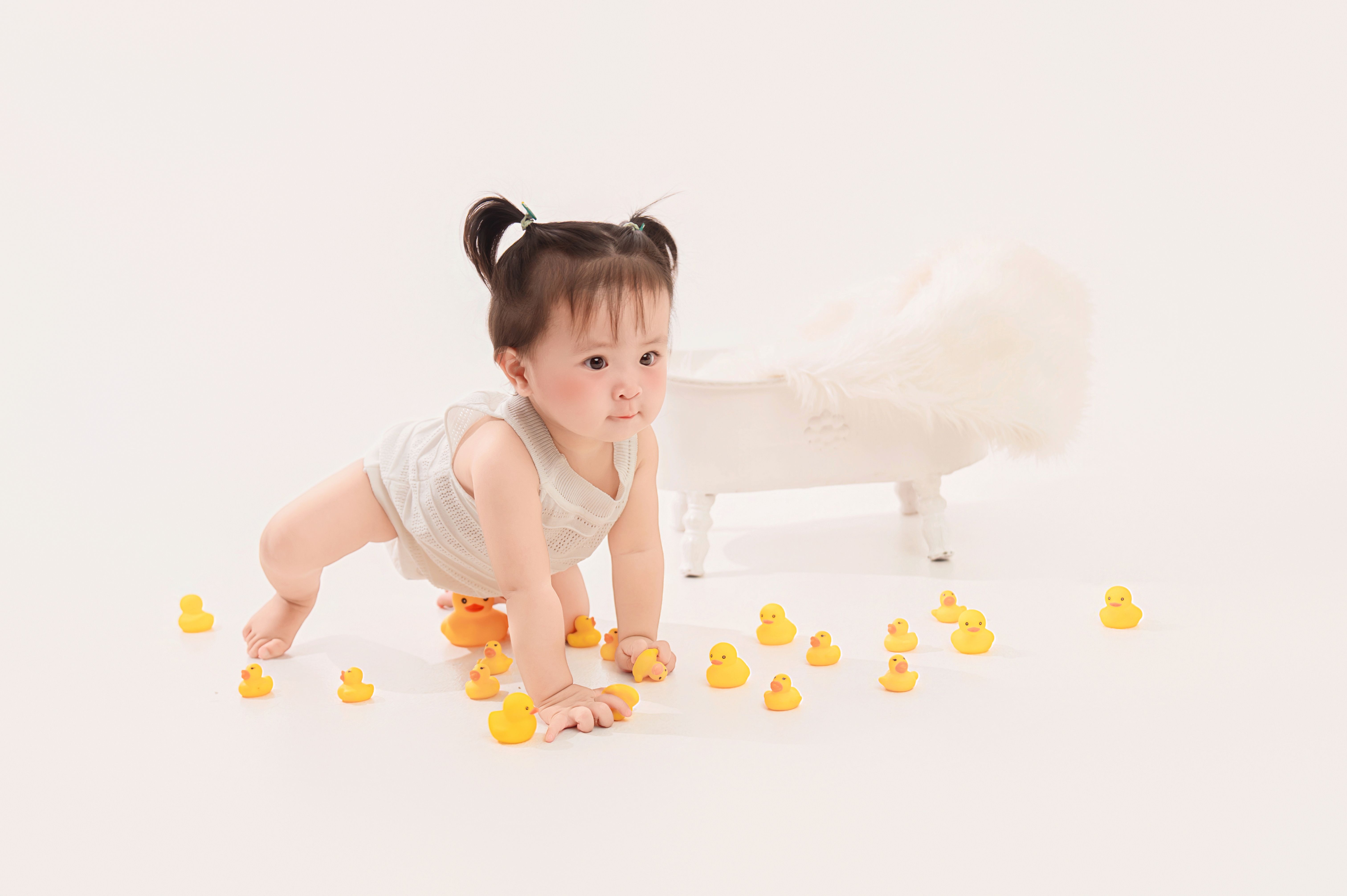 Cute baby with ponytails playing on the floor surrounded by yellow rubber ducks in a bright, minimalist setting.