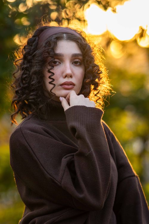 Free A thoughtful woman in a brown sweater, basking in warm sunset light against a blurry outdoor background. Stock Photo