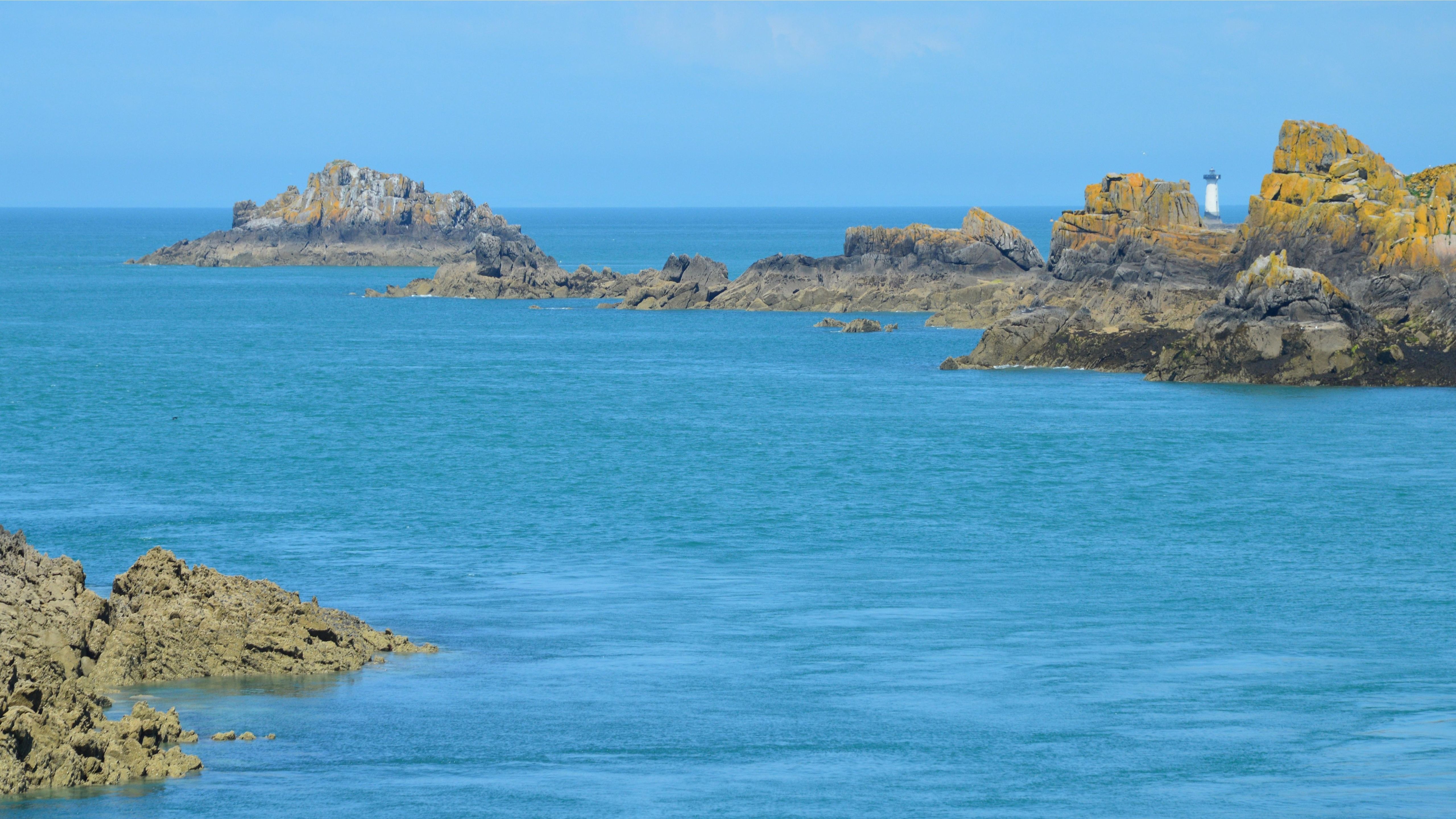 Explore the stunning rocky coastline in Cancale, Bretagne, France, with a distant lighthouse under a bright blue sky.