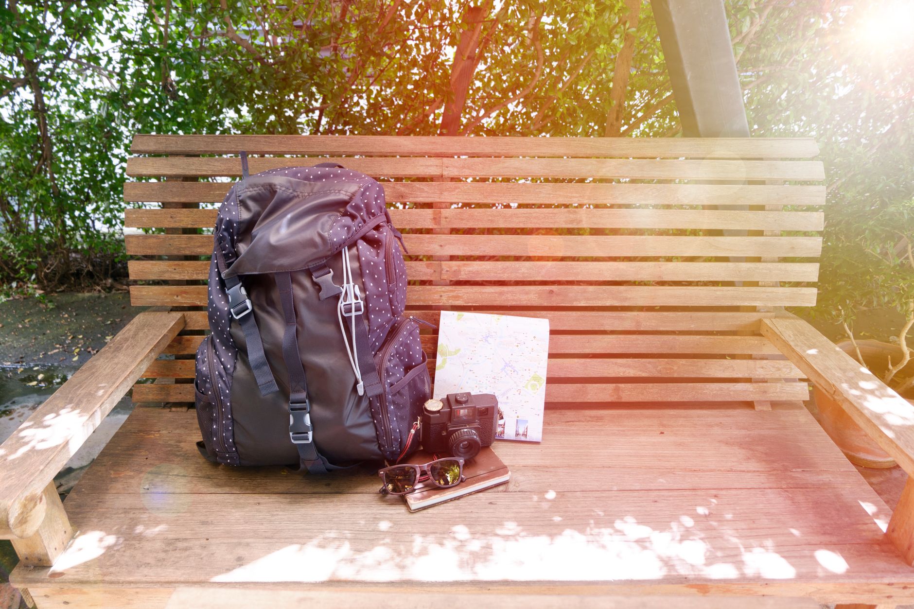 A backpack, camera, map, and sunglasses on a park bench with lens flare.