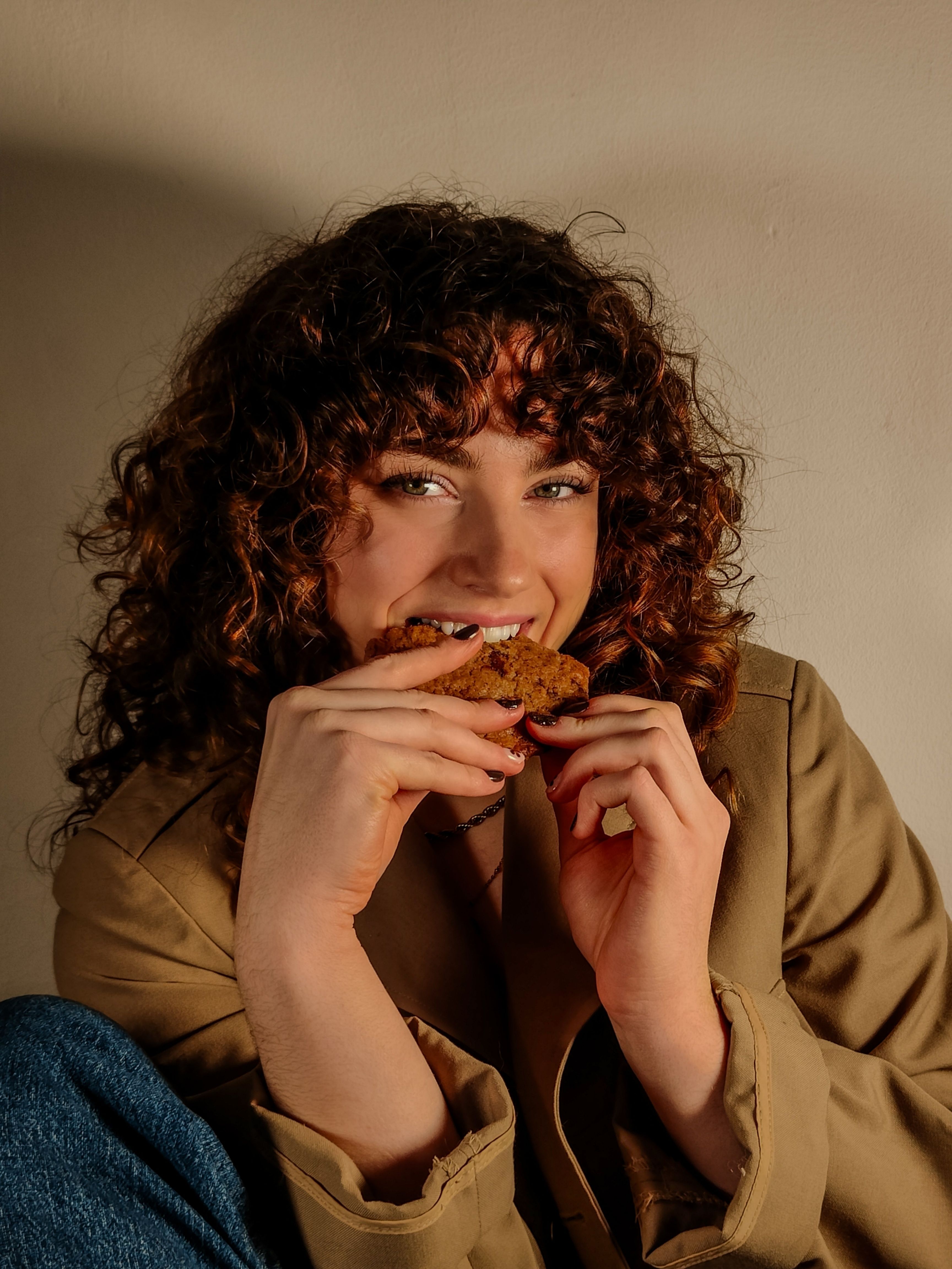Young woman with curly hair enjoying a cookie indoors. Relaxed and joyful moment.