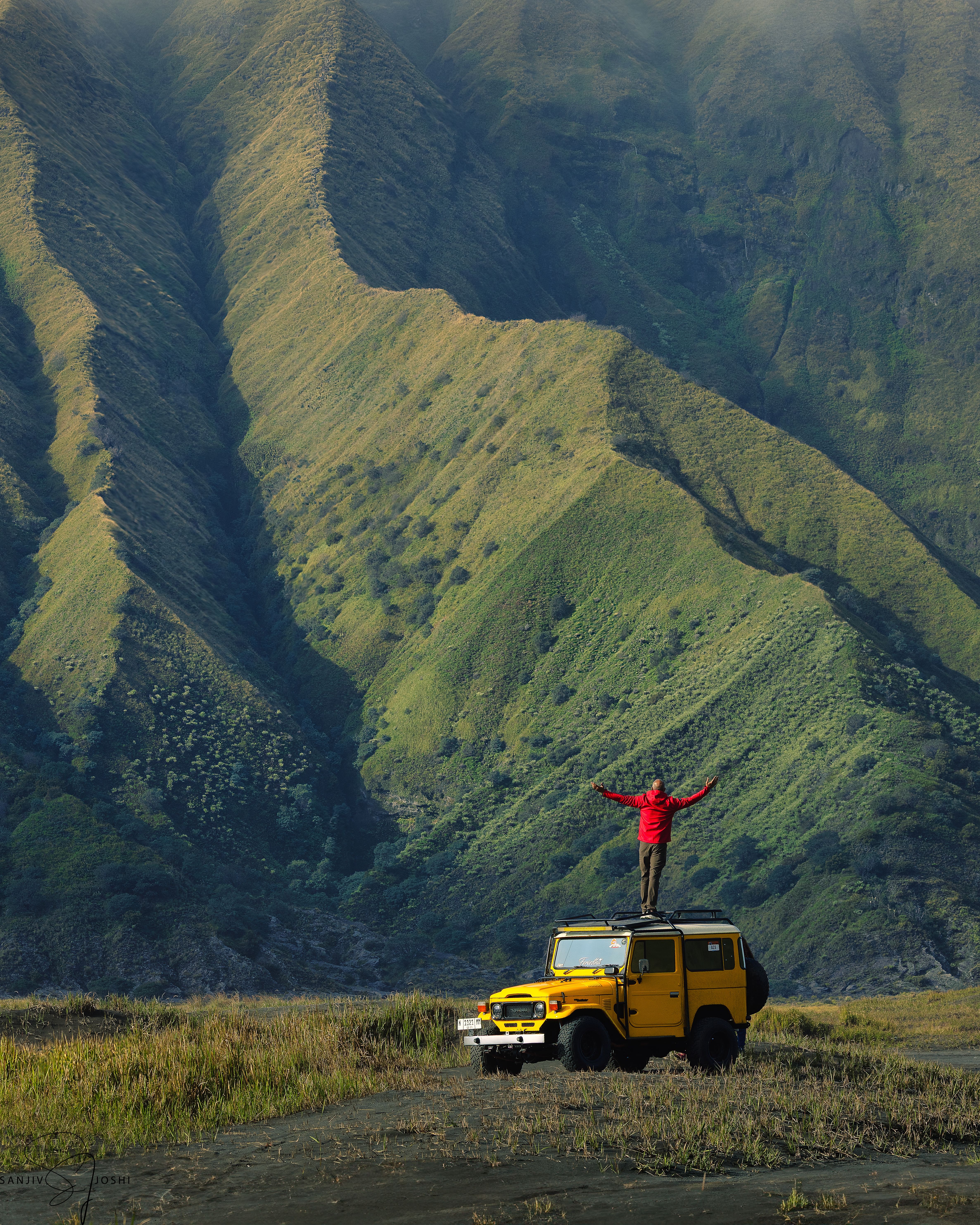 Fotomural Una persona aventurera de pie sobre un jeep amarillo con los brazos abiertos al pie de majestuosas montañas verdes.