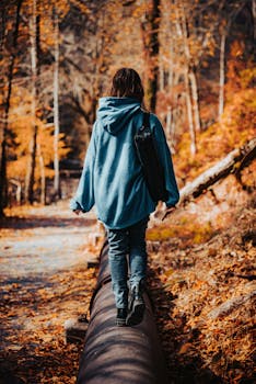 A young adult woman in a blue hoodie walks on a pipe through a vibrant autumn forest.