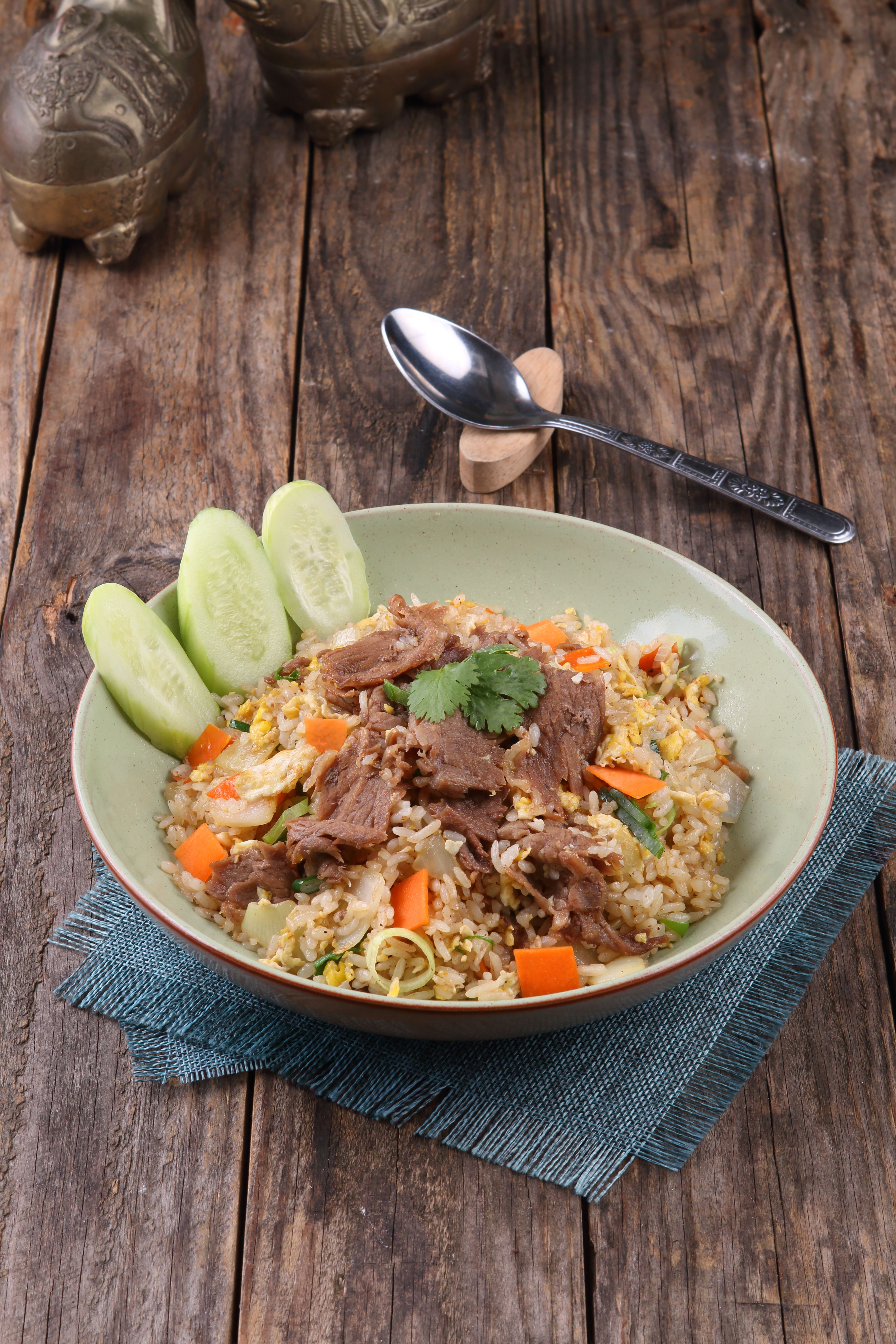 A vibrant bowl of beef fried rice with vegetables and cucumbers served on a rustic wooden table.
