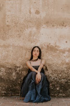 A woman in an elegant blue gown sits against a weathered vintage wall, exuding grace and serenity.