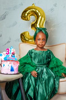 Young girl celebrating her third birthday with a cake and balloon indoors.