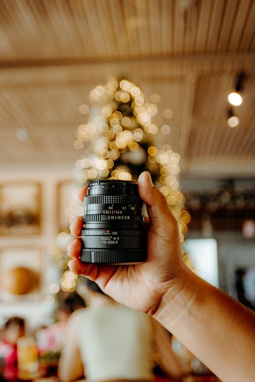 Free Close-up of a hand holding a camera lens in a warm, festive indoor setting with bokeh lights. Stock Photo