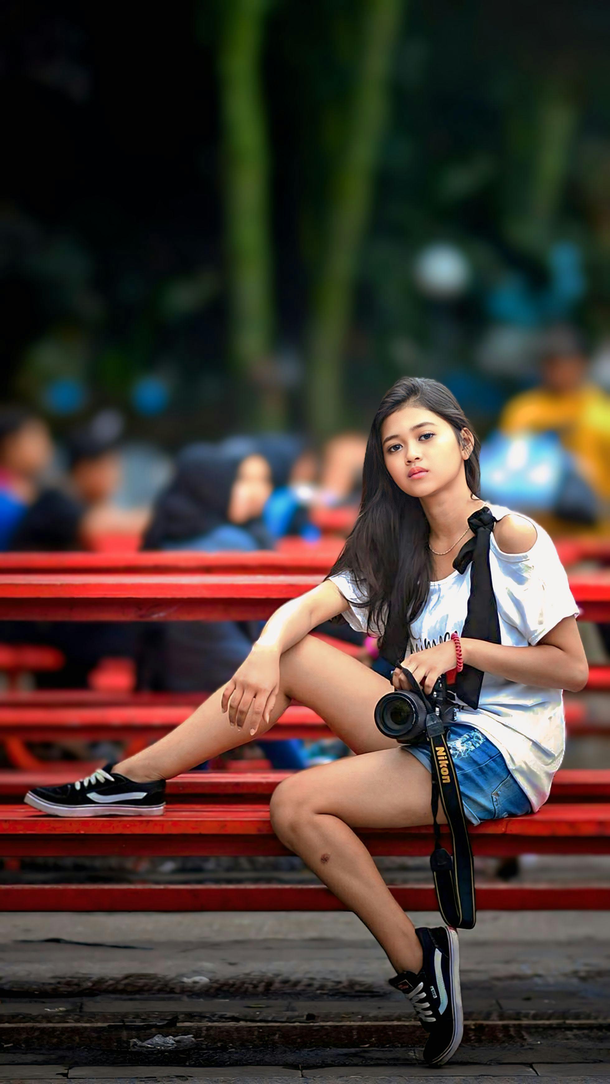 Casual young woman sitting on a red bench holding a camera, surrounded by people.