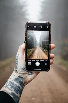 一只纹身的手拿着智能手机，拍下了一条薄雾笼罩的森林小路，展现了现代技术与自然的交融