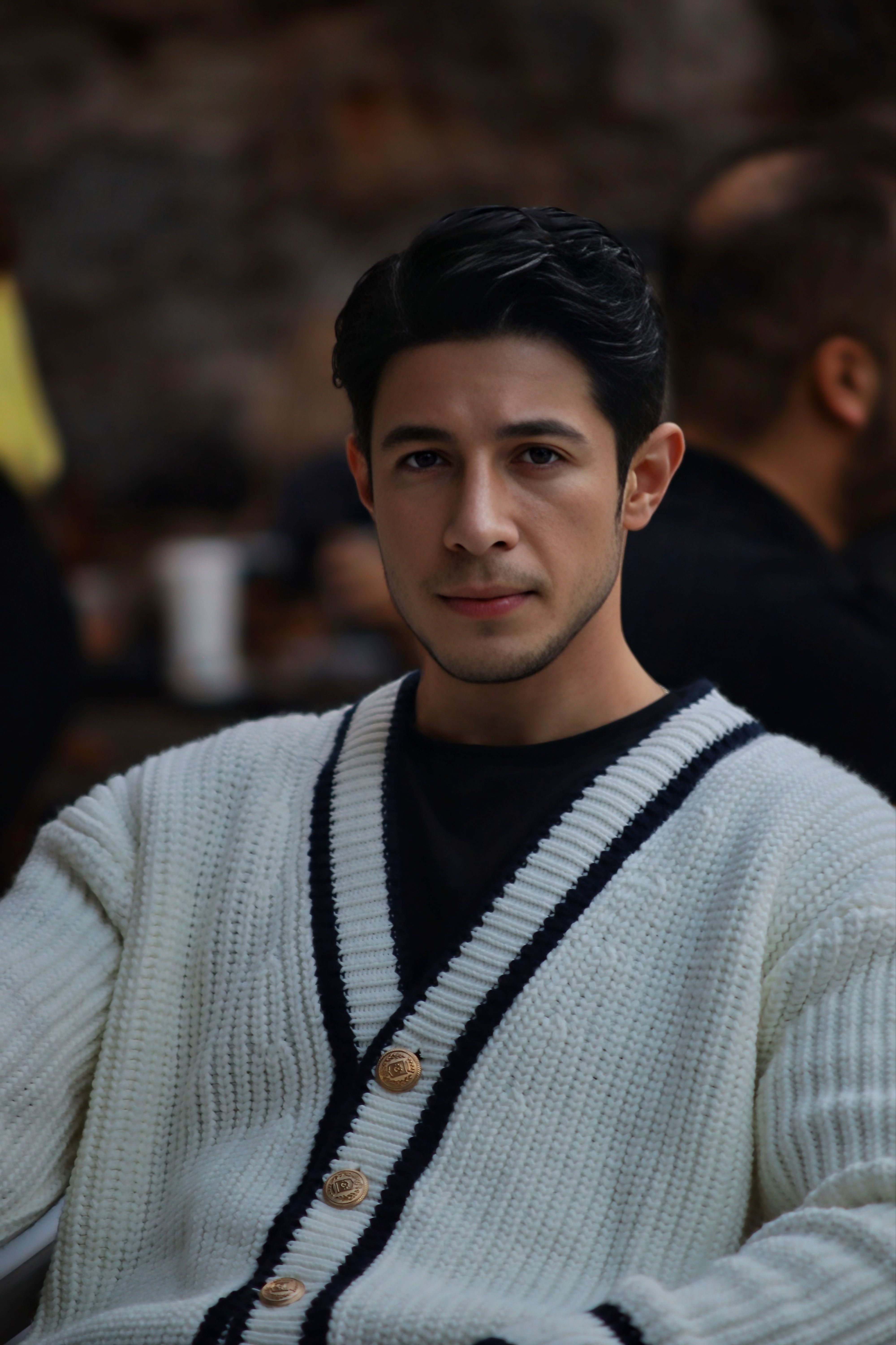 Man in an outdoor setting wearing a white and black cardigan, looking thoughtful.