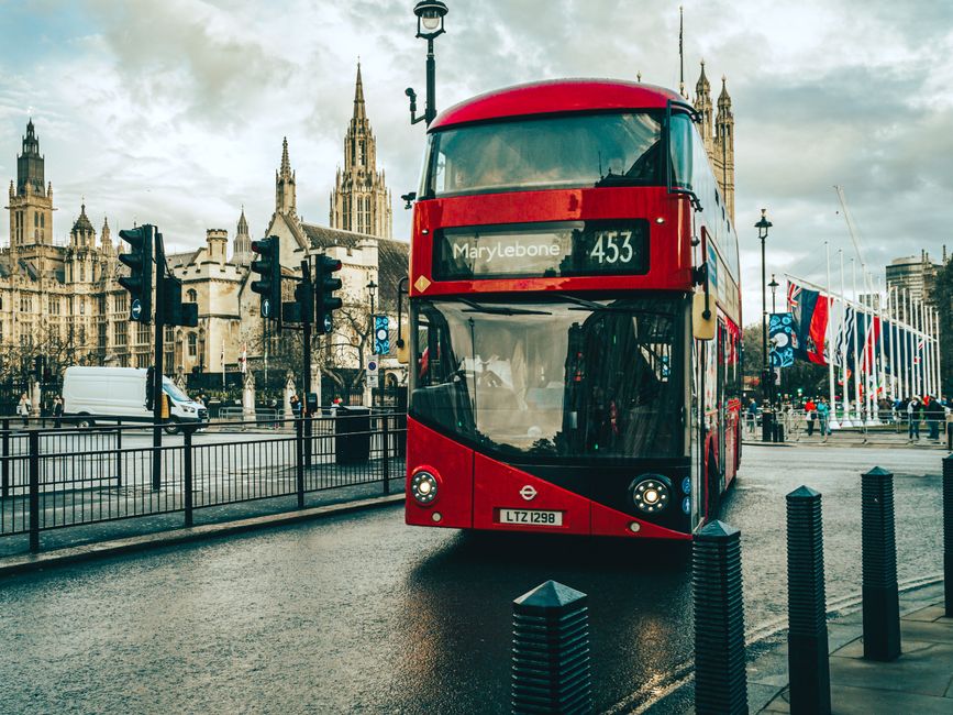 A classic red double-decker bus passing by famous landmarks in central London on a rainy day.