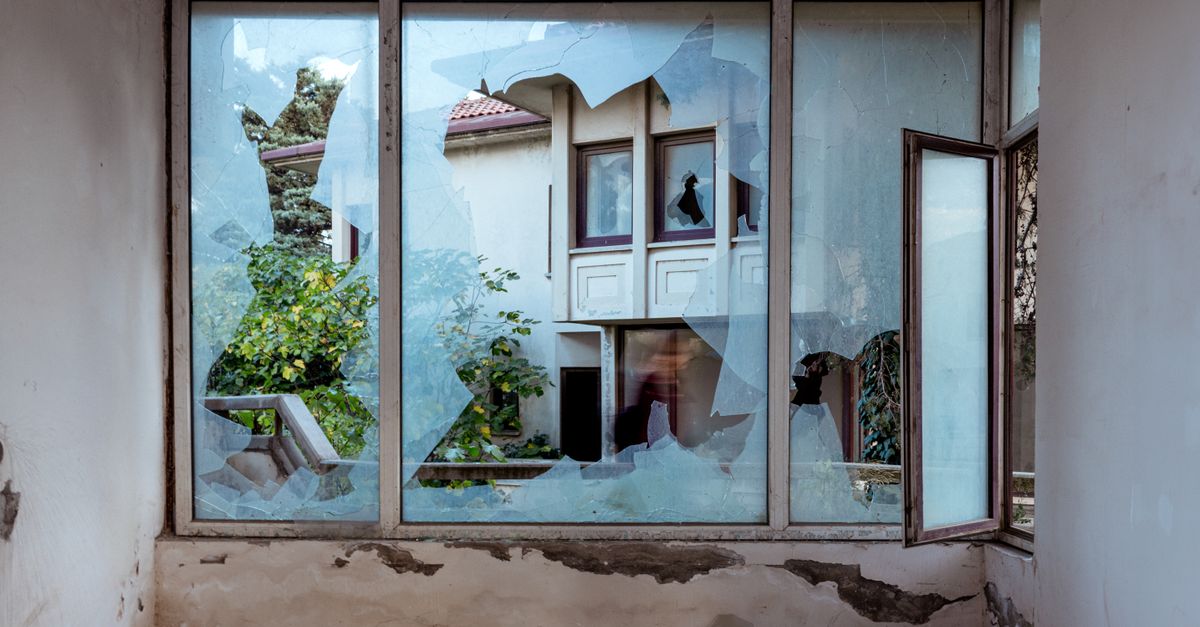 A shattered window revealing an abandoned building and greenery outside.