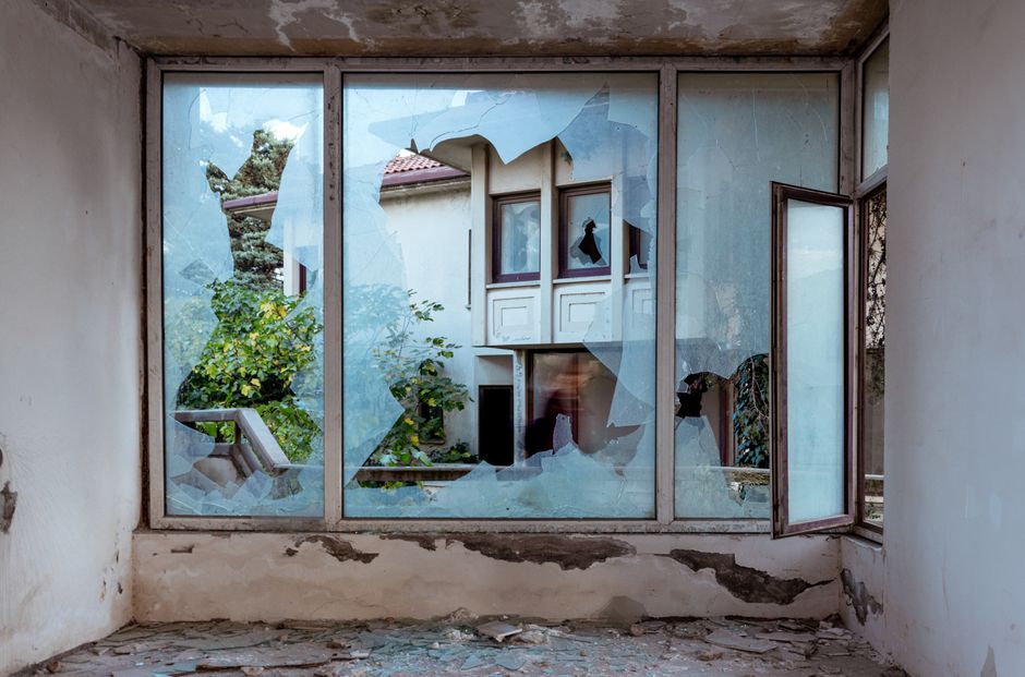 A shattered window revealing an abandoned building and greenery outside.