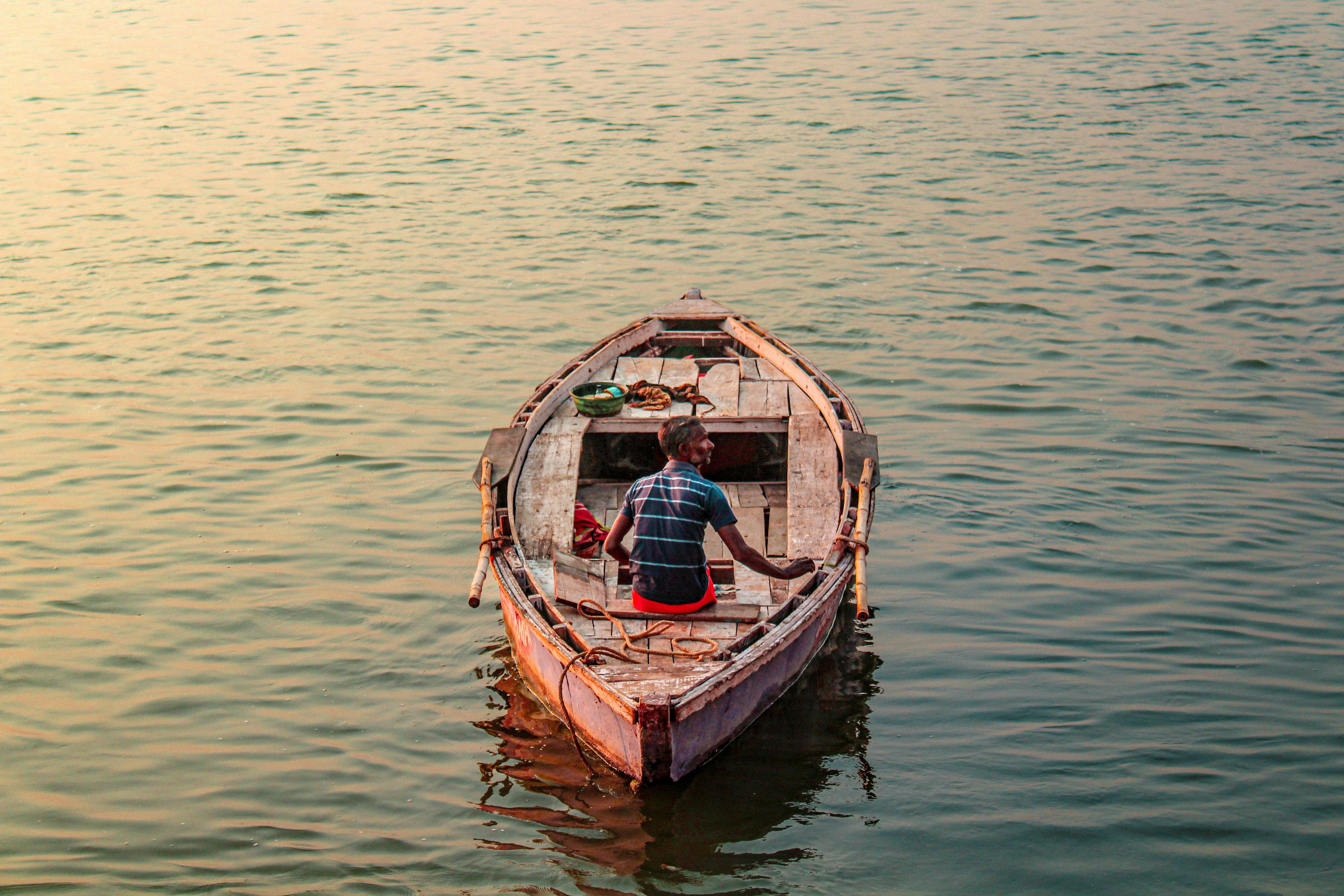 A lone fisherman sits on a wooden boat during sunset, casting a serene reflection.