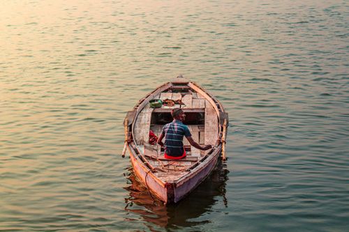 Free A lone fisherman sits on a wooden boat during sunset, casting a serene reflection. Stock Photo