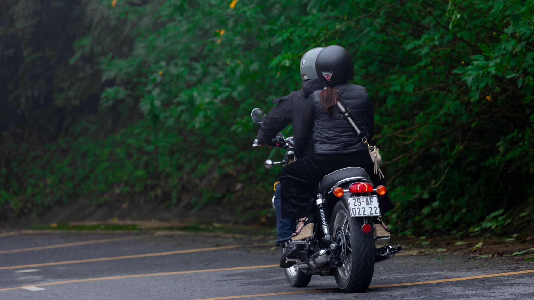 Two motorcyclists riding through a lush green road in Hanoi, Vietnam wearing helmets and protective gear.