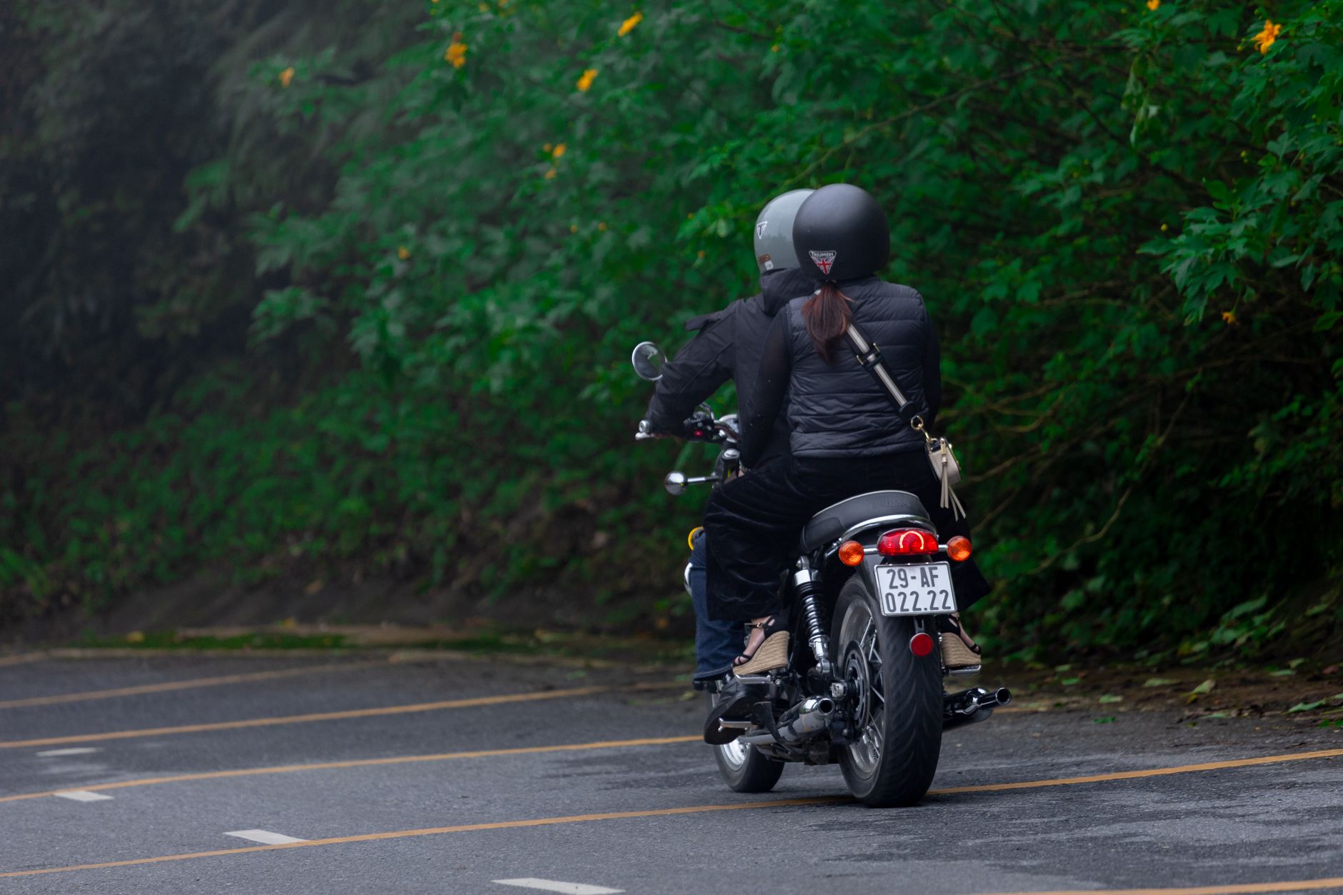 Two motorcyclists riding through a lush green road in Hanoi, Vietnam wearing helmets and protective gear.