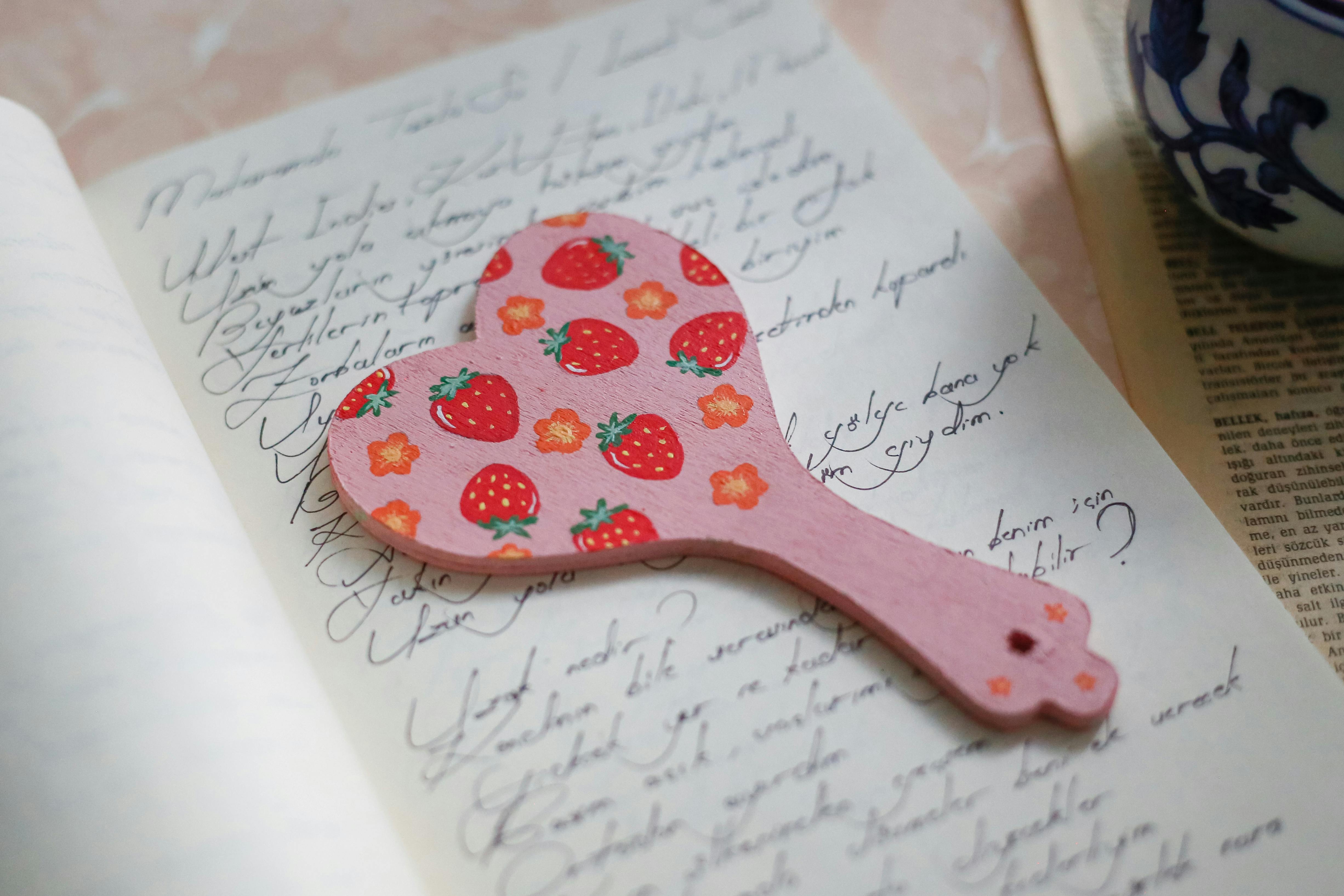A heart-shaped bookmark with strawberry design resting on handwritten notes.