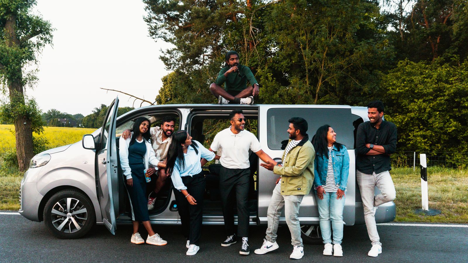 Eight adult friends smiling beside a passenger van during a relaxed road trip