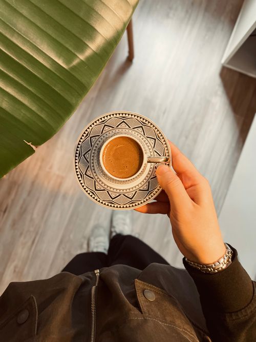 Free Person holding a patterned coffee cup indoors with plant leaf background, creating a cozy vibe. Stock Photo