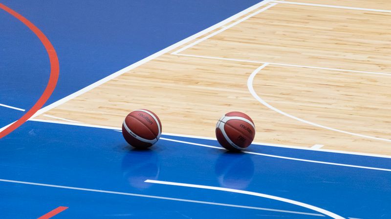 Two basketballs resting on a blue indoor basketball court with wooden flooring.