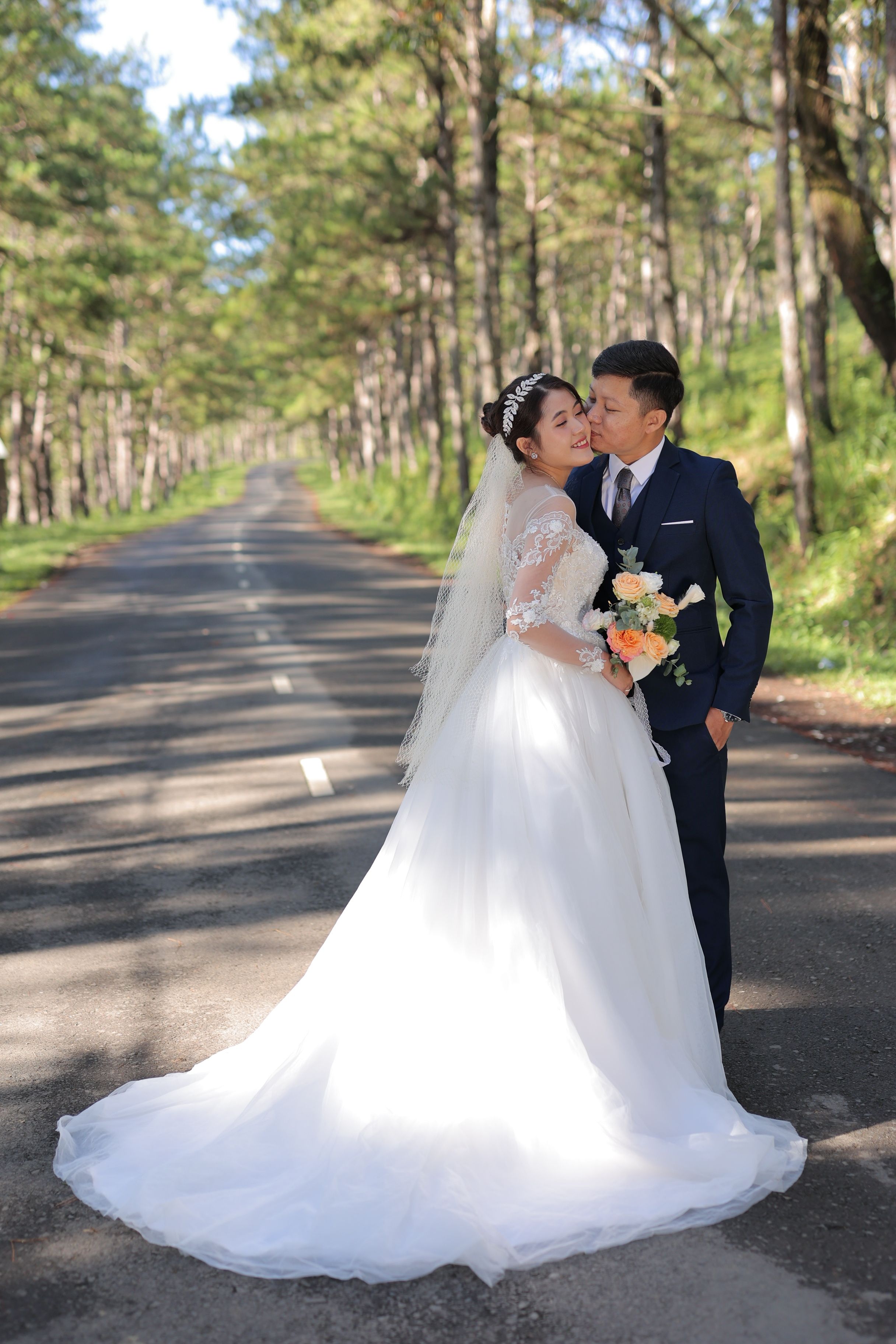 Bride and groom embrace on a sunlit forest road, capturing their special day with elegance.