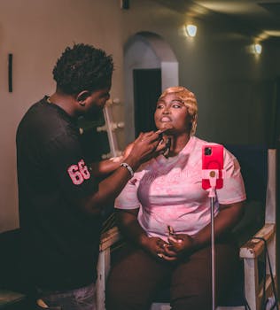 A makeup artist skillfully applies makeup indoors, captured in a warm, moody setting.