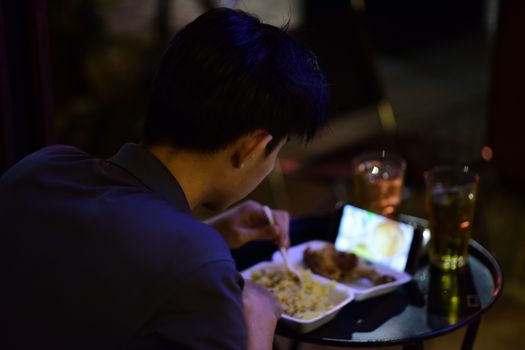 A young adult enjoys a meal while watching a video on their smartphone, creating a cozy night scene.