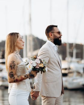Stylish couple posing by a marina, holding hands and flowers. Elegant and modern wedding scene.