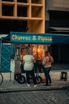 Street vendor stand offering churros and popcorn at night with two customers.