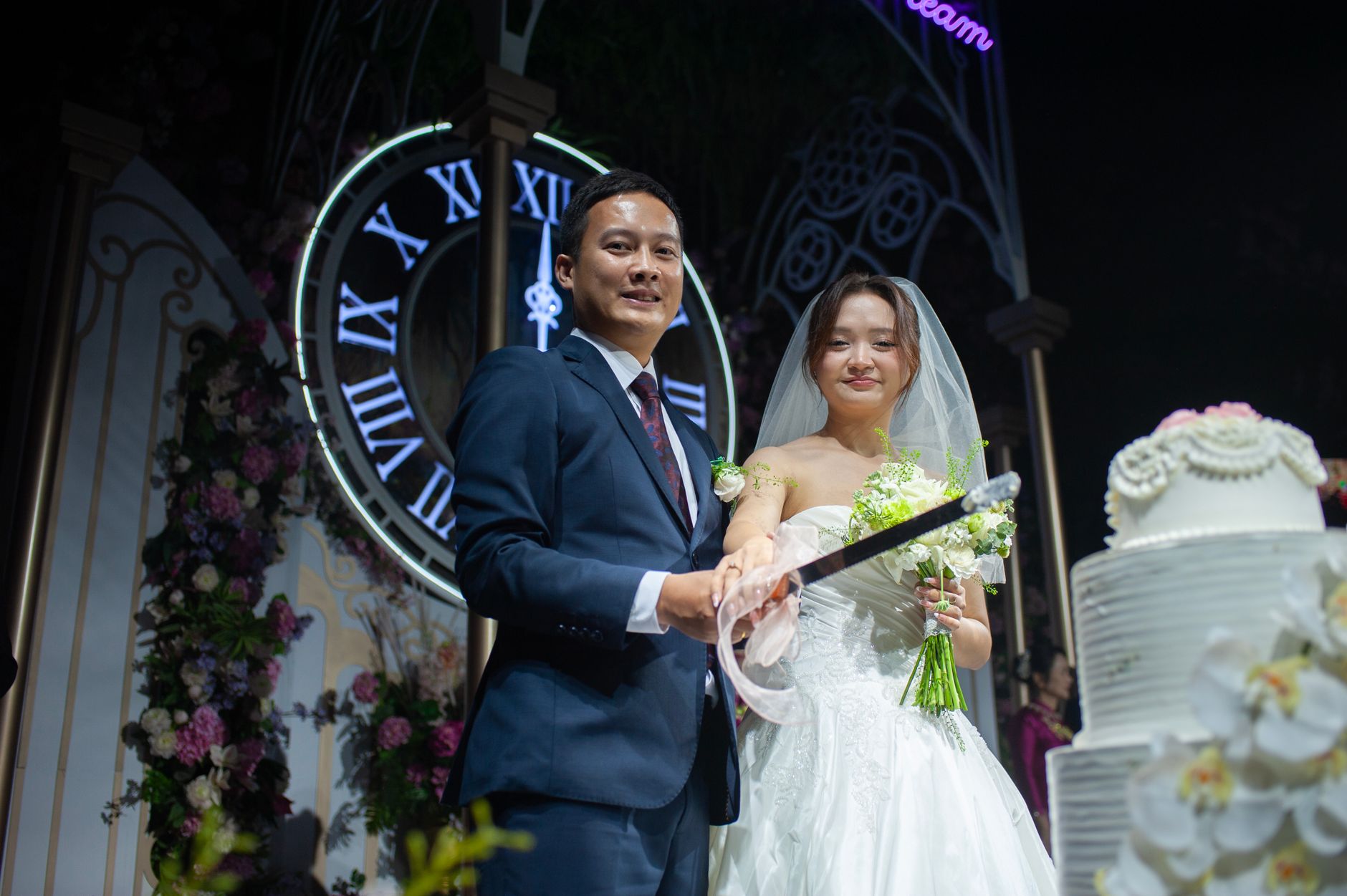 Bride and groom joyfully slicing wedding cake under ornate flower arch.