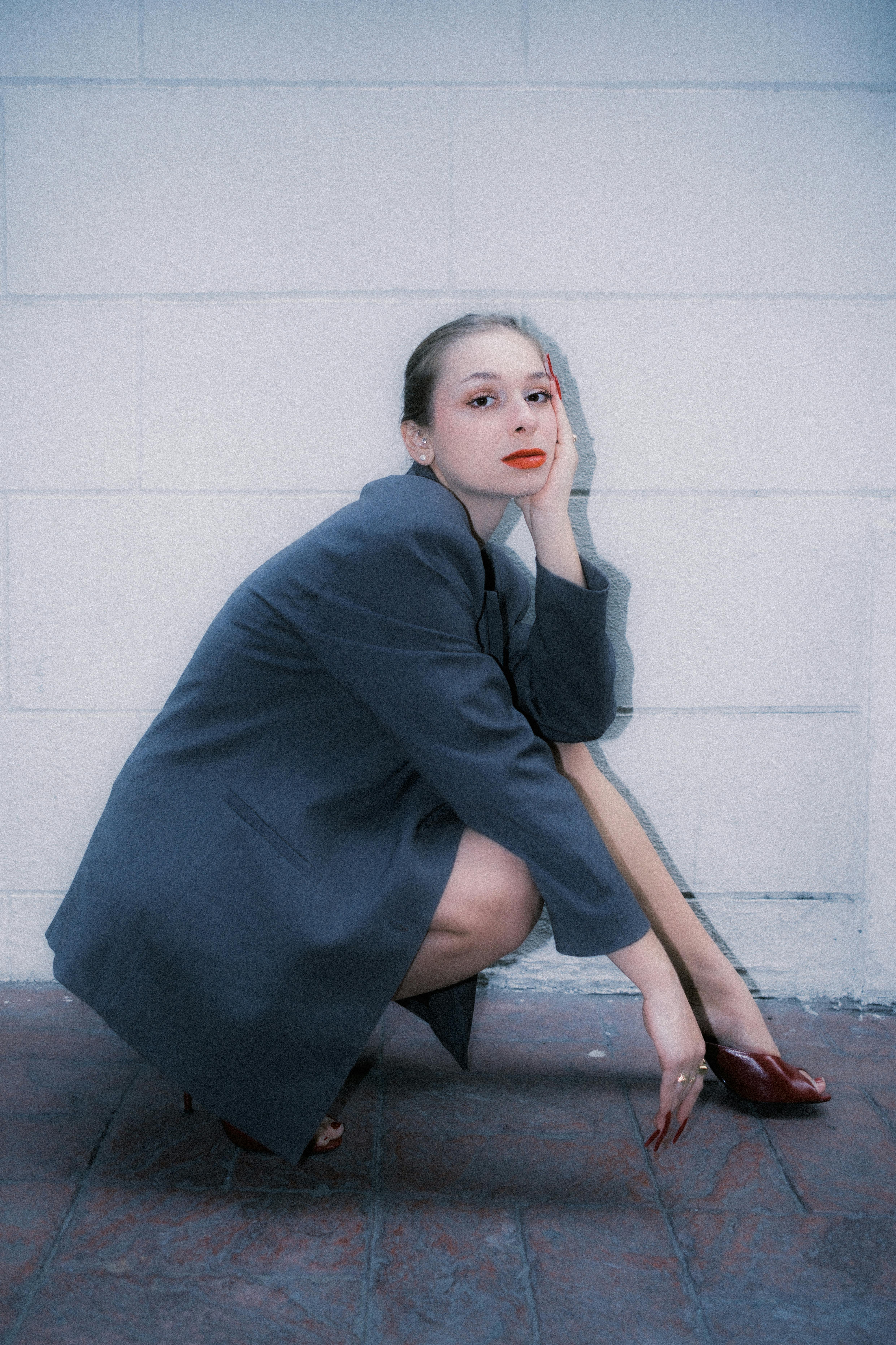 Kostenlos Eine Frau in einer grauen Jacke posiert elegant vor einer strukturierten weißen Backsteinwand in einem Innenraum. Stock-Foto