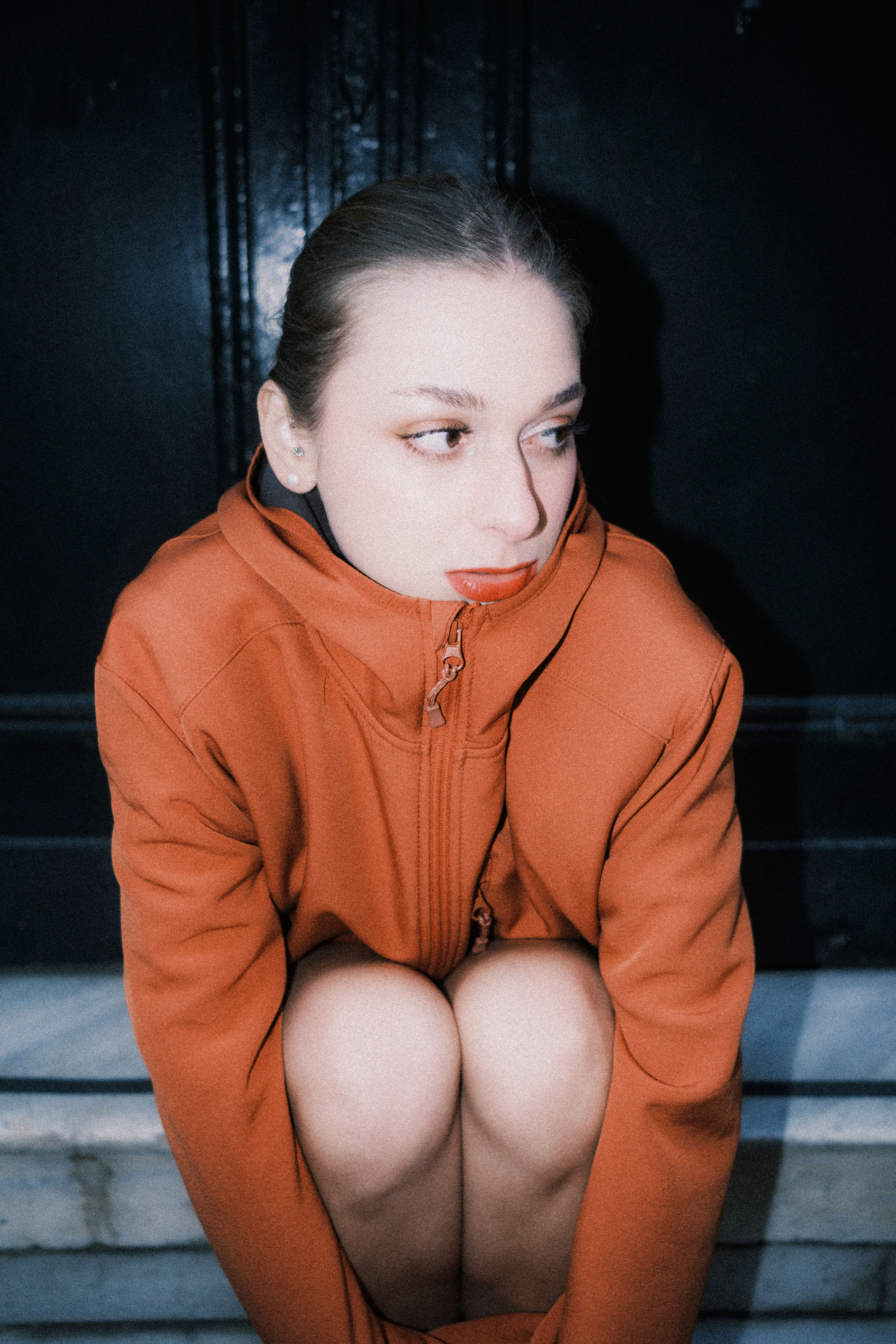 A contemplative woman in an orange jacket sits on steps indoors.
