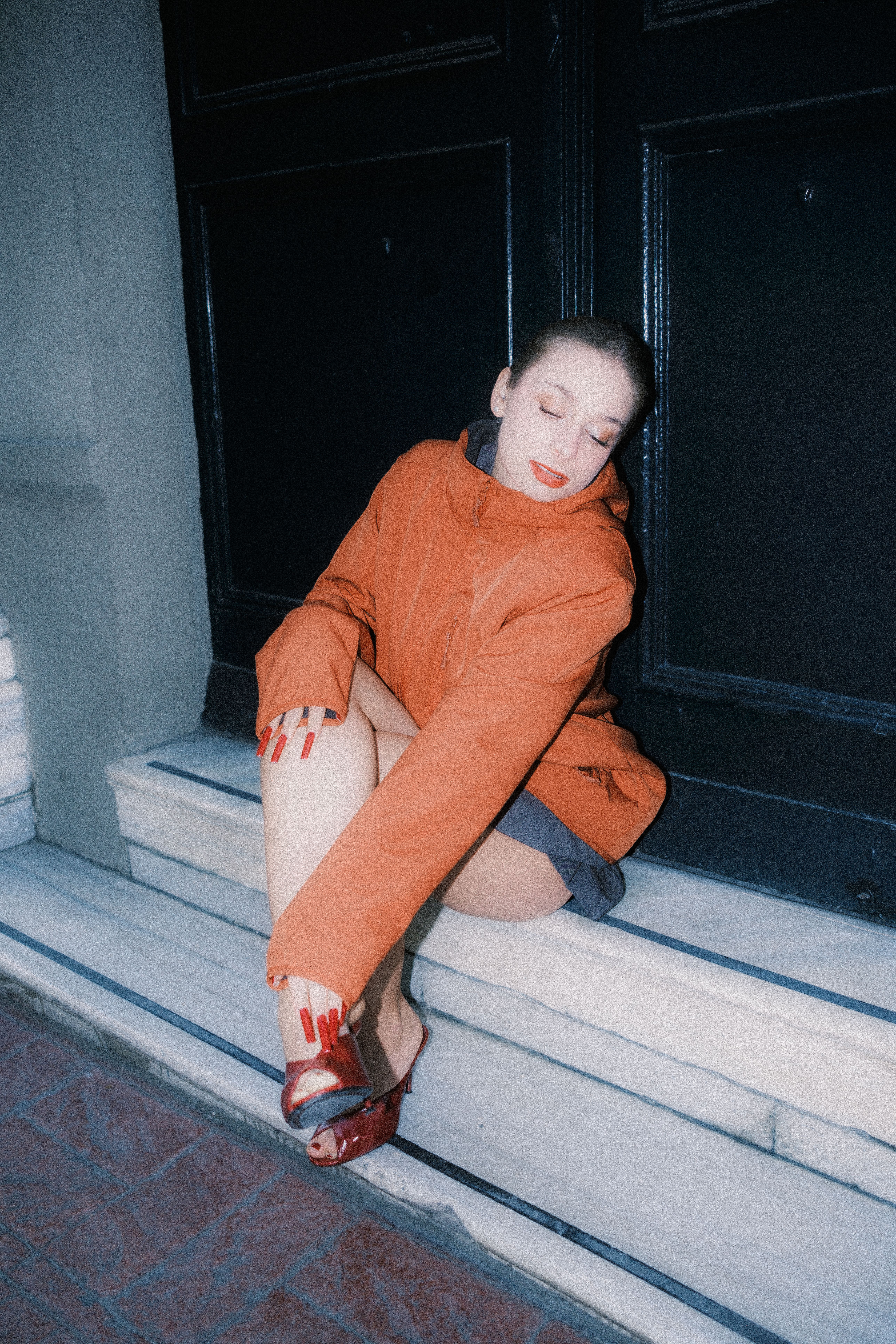 Stylish woman sitting on steps wearing an orange coat, exuding urban elegance.